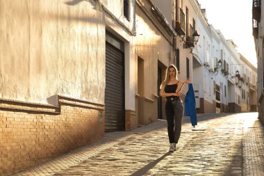 Young woman, beautiful and blonde, wearing a black tank top, blue fur jacket in her hand and jeans, walking down a lonely and lost street during the golden hour. Concept fashion, beauty, sunset.