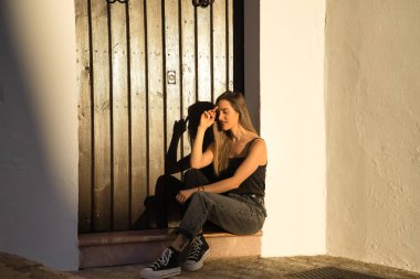 Young woman, beautiful and blonde, wearing a black tank top and jeans, sitting by a door, sad, alone and embarrassed. Concept sadness, grief, anxiety, loneliness.