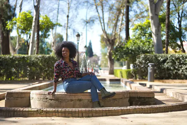 stock image Young woman, beautiful and black with afro hair, with flower shirt, jeans and boots, sitting in a fountain, very smiling and happy. Concept beauty, relaxation, smiles, happiness.