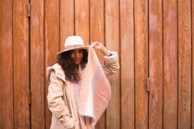 Genç, güzel, esmer, kıvırcık saçlı, paltolu, atkısı ve şapkası olan, eşarbıyla komik bir şekilde oynarken kameraya bakan, ahşap arka planda. Güzellik, moda, sonbahar, kış, soğuk kavramları.