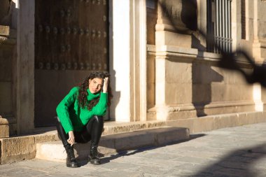 Genç, güzel, kıvırcık saçlı, yeşil boğazlı kazaklı, deri pantolonlu, siyah çizmeli mutlu ve rahat bir şekilde merdivende oturan esmer bir kadın. Güzellik, moda, sonbahar, kış, soğuk kavramları.