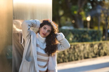 Genç, güzel, kıvırcık saçlı, kazaklı, ceketli ve güneş gözlüklü, metal bir duvara yaslanmış, şehvetli ve cilveli, güneş ışınlarını alan bir kadın. Güzellik, moda, sonbahar, kış, soğuk kavramları