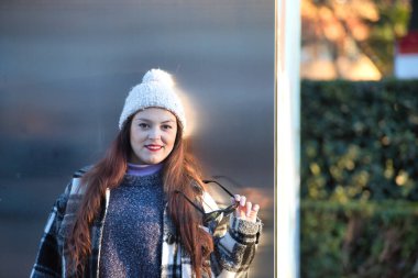 Güzel, düz kahverengi saçlı, kazak, ceket, yün şapka ve güneş gözlüklü, metal bir duvara yaslanmış, cilveli ve şehvetli bir kadın. Güzellik, moda, sonbahar, kış, soğuk kavramları.