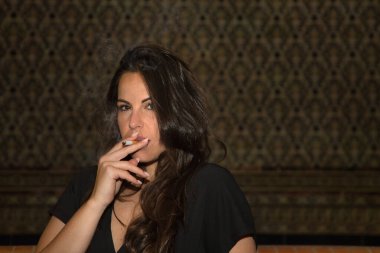 Genç ve güzel bir kadın, İspanyol, kahverengi saçlı, siyah bir elbise giymiş, merdivenlerde oturmuş sigara içiyor, kameraya şehvetli ve kışkırtıcı bakıyor. Kavram tütün, bağımlılık, sağlık.