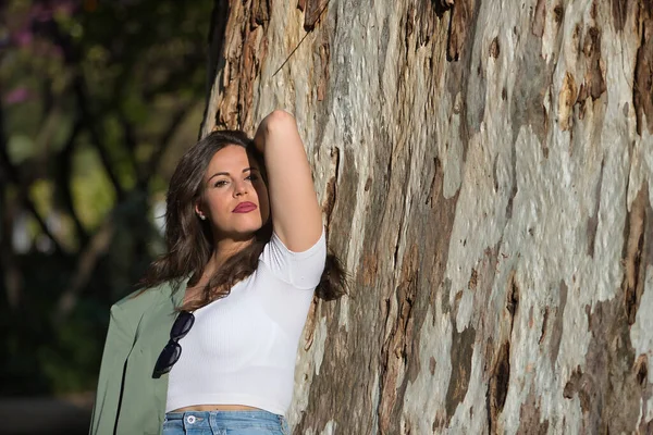 年轻而美丽的女人 西班牙裔 棕色的头发 穿着绿色的夹克和牛仔裤 看着相机 一个人 放松而镇定 靠着一棵大树的树干 镇定的概念 — 图库照片