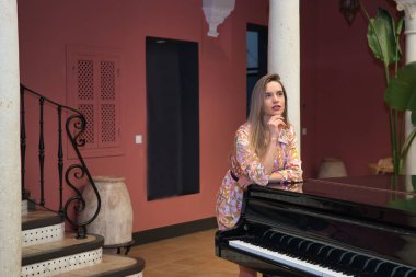 Çiçekli elbiseli, genç, güzel, sarışın bir kadın, düşünceli, güzel bir piyanonun üzerine yaslanmış, lüks bir otelin resepsiyonunda. Kavram araçları, düşünceler, binalar, iç tasarım.