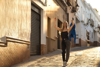 Young woman, beautiful and blonde, wearing a black tank top, blue fur jacket in her hand and jeans, walking down a lonely and lost street during the golden hour. Concept fashion, beauty, sunset.