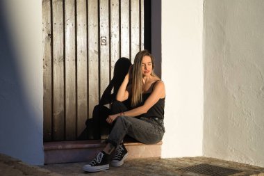 Young woman, beautiful and blonde, wearing a black tank top and jeans, sitting by a door, sad, alone and embarrassed. Concept sadness, grief, anxiety, loneliness.