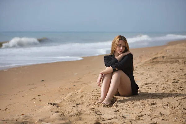 Young, blonde, beautiful woman in a bikini and black colored shirt sitting on the beach, sad, lonely, sorry, depressed. Concept loneliness, sadness, grief, depression.