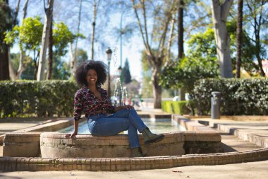 Genç bir kadın, güzel ve siyah, afro saçlı, çiçekli gömlekli, kot pantolonlu ve botlu, çeşmede oturuyor, çok mutlu ve gülümsüyor. Güzelliği kavra, rahatlamayı, gülümsemeyi, mutluluğu..