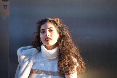 Genç, güzel, kıvırcık saçlı, kazaklı, ceketli ve güneş gözlüklü, metal bir duvara yaslanmış, şehvetli ve cilveli, güneş ışınlarını alan bir kadın. Güzellik, moda, sonbahar, kış, soğuk kavramları