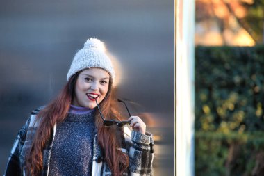 Young woman, beautiful, straight brown hair, sweater, coat, wool hat and sunglasses, leaning on a metal wall, playing with her glasses, flirtatious and sensual. Concept beauty, autumn, winter, cold.