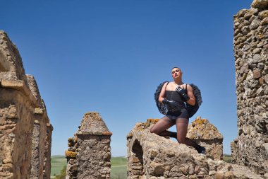 İkili olmayan, genç ve Güney Amerikalı, çok makyajlı, siyah kanatlı ve deri eldivenli, korse ve siyah çoraplı, diz çökmüş, bir ortaçağ kalesinin yanında poz veriyor..