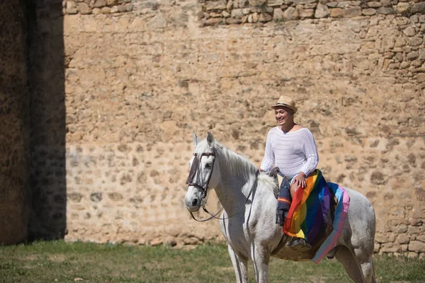 stock image Non-binary person, young and South American, very makeup, mounted on a white horse, smiling and happy, with a gay pride flag on the rump, next to an old medieval castle. Concept queen, lgbtq+, pride.