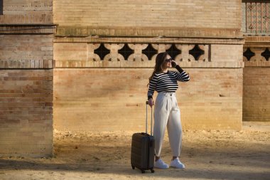 Genç ve güzel bir kadın, İspanyol, kahverengi saçlı, çizgili kazaklı, keten pantolonlu ve güneş gözlüklü, gün batımında cep telefonunun yanında bekliyor, konuşuyor. Seyahat, tatil, bavul