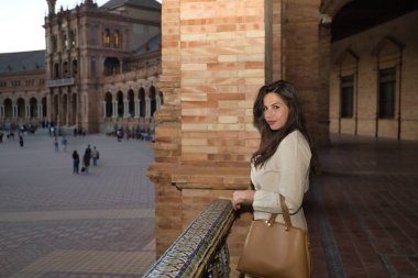 Genç ve güzel bir kadın, İspanyol, kahverengi saçlı, açık renk ceketli ve kahverengi çantalı, fayanslı korkuluklara yaslanmış, kameraya bakıyor, alacakaranlıkta rahat ve sakin. Güzellik ve moda kavramı.