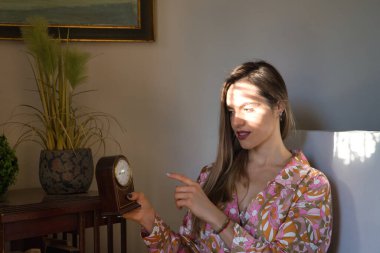 Young, pretty, blonde woman in a flowered dress, sitting, pointing to an antique clock and receiving sun rays on her face through a shutter. Concept time, clocks, waiting, sunbeams.