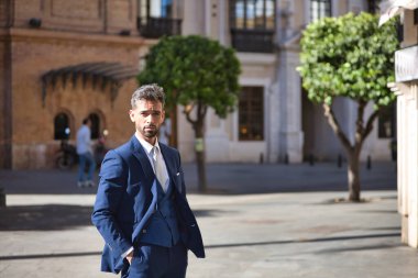 Çekici genç iş adamı, sakallı ve takım elbiseli, elleri cebinde şehrin ortasında duruyor. Güzelliği, modayı, başarıyı, başarıyı, eğilimi kavra.