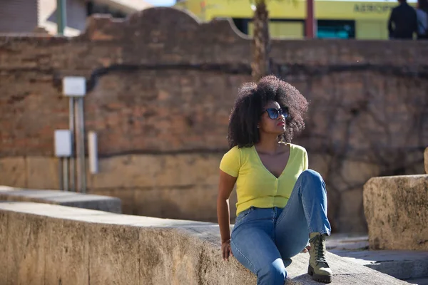 美しい アフロの髪を持つ黒の女性は 黄色のTシャツ ジーンズやサングラスを着て 石の壁に座って リラックスし 穏やかな日光浴 休暇の概念 — ストック写真