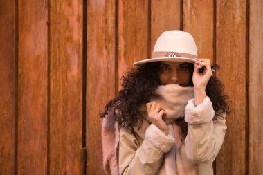 Young, beautiful, brunette woman with curly hair and coat, scarf and hat, looking at camera while protecting herself from cold with scarf, on wooden background. Concept fashion, autumn, winter, cold.