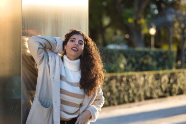 Genç, güzel, kıvırcık saçlı, kazaklı, ceketli ve güneş gözlüklü, metal bir duvara yaslanmış, şehvetli ve cilveli, güneş ışınlarını alan bir kadın. Güzellik, moda, sonbahar, kış, soğuk kavramları