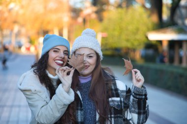 İki kadın, genç ve güzel, kazak, ceket ve yün şapkalar içinde, gülümseyen ve mutlu, gözlerini kuru yapraklarla kapatıyorlar. Sonbaharı, kışı, soğuğu, arkadaşlığı düşün. Kuru yapraklara odaklan.
