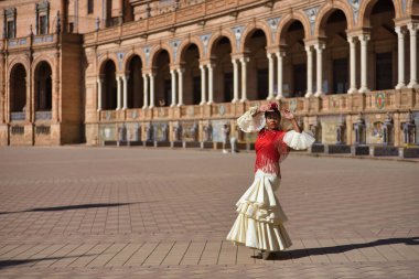 Bej renkli çingene flamenko takım elbiseli ve kırmızı şallı genç siyah ve Güney Amerikalı kadın İspanya 'nın Seville şehrinde güzel bir meydanda dans ediyor. Konsept dansı, folklor, flamenko, sanat.