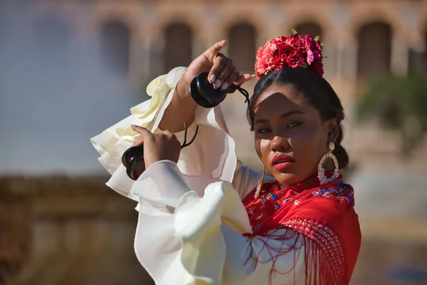 Stock image Young black and South American woman in a beige gypsy flamenco suit and red shawl, dancing with castanets in a beautiful square in the city of Seville in Spain. Concept dance, folklore, flamenco, art