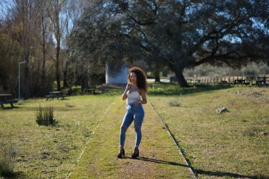 Genç, güzel, esmer, kıvırcık saçlı, beyaz bluzlu, kot ve topuklu giyen, parkın ortasında kameraya bakarak poz veren bir kadın. Güzelliği, modayı, pozu kavra.