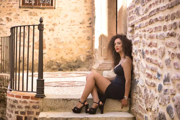 Genç bir kadın, güzel, esmer, kıvırcık saçlı, siyah elbiseli ve topuklu ayakkabılı, kaybolmuş bakışlı, üzgün ve yalnız, taş basamaklarda oturuyor. Moda, güzellik, model, üzüntü, yalnızlık.