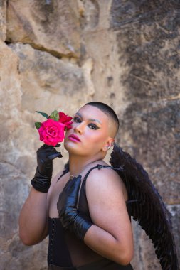 İkili olmayan birinin portresi, genç ve Güney Amerikalı, çok makyajlı, siyah kanatlı ve deri eldivenli, korse, yüzüne güller yapışmış şekilde poz veriyor. Kavram kraliçesi, lgbtq +. Dikey konum.