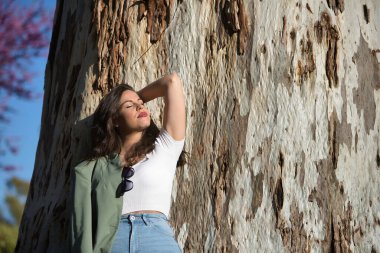 Genç ve güzel bir kadın, İspanyol, kahverengi saçlı, yeşil ceketli ve kotlu, gözleri kapalı, yalnız, rahat ve sakin, kocaman bir ağacın gövdesine yaslanmış. Yalnızlık kavramı, rahatla, sakinleş..