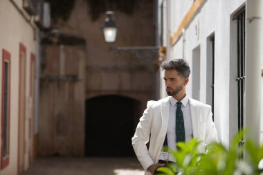 Sakallı, takım elbiseli ve kravatlı çekici genç işadamı, yeşil bitkilerin arkasında elleri ceplerinde poz veriyor. Güzelliği, modayı, başarıyı, başarıyı, eğilimi kavra.