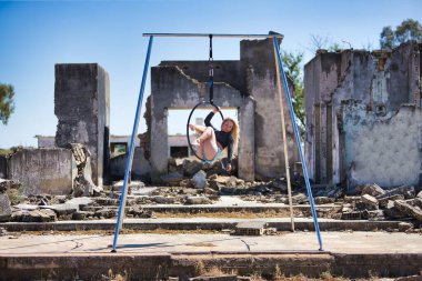 Genç, sarışın, atletik bir kadın. Havada asılı duran bir halkayla bazı yıkık binaların yanında egzersiz yapıyor. Kavramsal çember, yoga ve direk dansı.