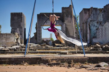 Genç bir kadın, sarışın ve atletik. Havada asılı duran bir halkayla baş aşağı egzersiz yapıyor. Rüzgarda savrulan beyaz bir duvak, harabeler içindeki bazı binaların yanında..