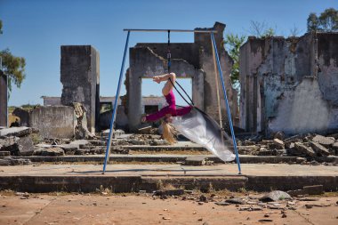 Genç bir kadın, sarışın ve atletik. Havada asılı duran bir halkayla baş aşağı egzersiz yapıyor. Rüzgarda savrulan beyaz bir duvak, harabeler içindeki bazı binaların yanında..