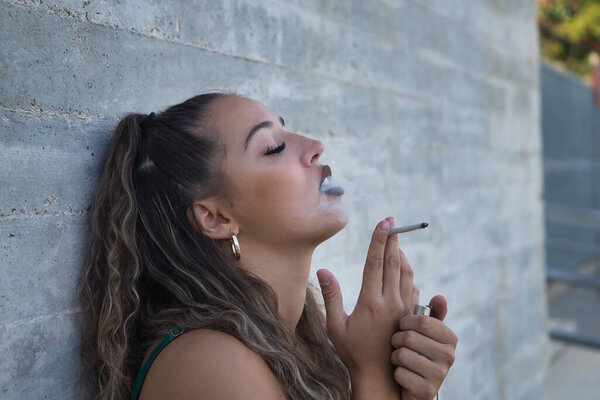 Латиноамериканка и латиноамериканка, молодая и нонконформистка, бунтарка, курит сигарету, выдыхая дым чувственным способом, на сером фоне. Табак, дым, огонь, зависимость