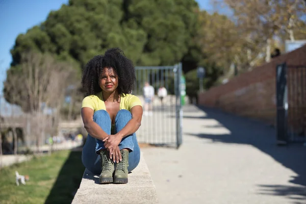 Young, beautiful, black woman with afro hair, wearing yellow t-shirt, jeans and boots, sitting on a stone wall, clutching her knees, calm and relaxed. Concept relax, tranquility, peace.