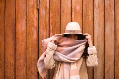 Genç, güzel, esmer, kıvırcık saçlı, paltolu, atkısı ve şapkası olan, eşarbıyla komik bir şekilde oynarken kameraya bakan, ahşap arka planda. Güzellik, moda, sonbahar, kış, soğuk kavramları.