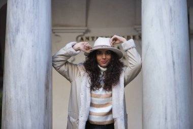 Genç, güzel, esmer, kıvırcık saçlı, bej kazaklı, palto ve şapkalı, mermer kolonlara yaslanmış, mutlu ve rahat. Güzellik, moda, sonbahar, kış, soğuk kavramları.