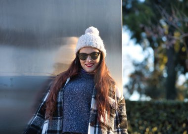 Güzel, düz kahverengi saçlı, süveter, ceket, yün şapka ve güneş gözlüklü, metal bir duvara yaslanmış, gülümseyen ve mutlu bir kadın. Güzellik, moda, sonbahar, kış, soğuk kavramları.