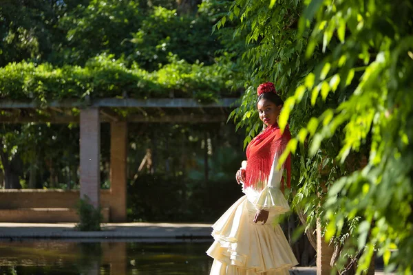 stock image Young black and South American woman in a beige gypsy flamenco suit and red shawl, posing in a park in the city of Seville in Spain. Concept dance, folklore, flamenco, art.