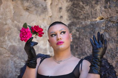 İkili olmayan bir kişinin portresi, genç ve Güney Amerikalı, çok makyajlı, siyah kanatlı ve deri eldivenli, korse, elinde güllerle poz veriyor. Kavram kraliçesi, lgbtq +, gurur, nonoş.