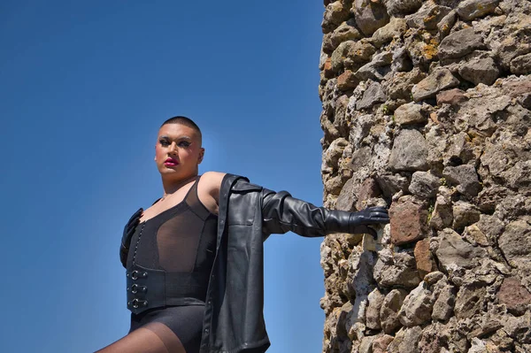 İkili olmayan bir kişinin portresi, genç ve Güney Amerikalı, makyajlı, siyah ceketli, deri eldivenli, korse ve siyah çoraplı, ortaçağ şatosunun kalesinin yanında poz veren.