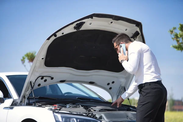 Young hispanic man, wearing a white shirt, calling the tow truck with his mobile, after suffering a breakdown in his white luxury car. Concept, cars, breakdowns, roadside assistance, crane.