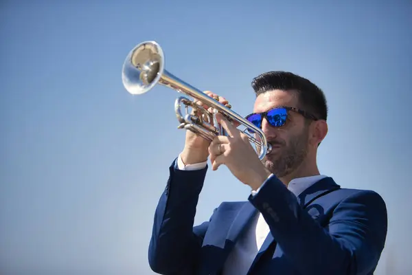 Young Hispanic man, wearing a jacket and sunglasses, playing a pretty, silvery trumpet outdoors. Concept, music, instruments, trumpet.