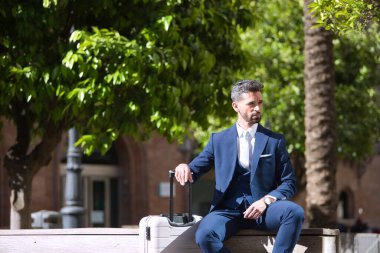 Sakallı ve takım elbiseli genç ve çekici bir işadamı, bavulunun yanındaki bankta oturmuş taksi bekliyor. Güzelliği, modayı, başarıyı, başarıyı, eğilimi kavra.