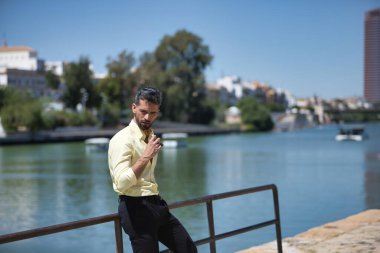 Sakallı, sarı gömlekli, siyah pantolonlu, güneş gözlüklü ve korkuluğa yaslanmış genç ve çekici bir iş adamı. Güzelliği, modayı, başarıyı, başarıyı, eğilimi kavra.