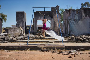 Genç bir kadın, sarışın ve atletik. Havada asılı duran bir halkayla baş aşağı egzersiz yapıyor. Rüzgarda savrulan beyaz bir duvak, harabeler içindeki bazı binaların yanında..
