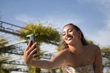 Latin ve İspanyol kız, genç ve güzel, cep telefonuyla video kaydediyor, komik hareketler yapıyor. Konsept cep telefonu, akıllı telefon, fotoğraf, video, uygulamalar, kayıt.